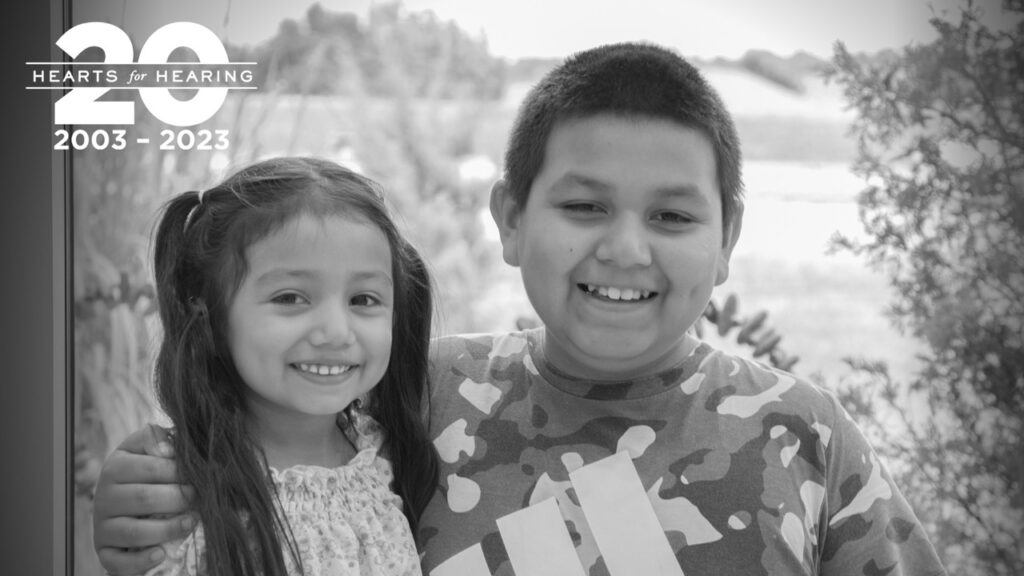Two kids stand side by side hugging. The girl on the left has pigtails. The boy on the right has short hair. Both have large smiles.