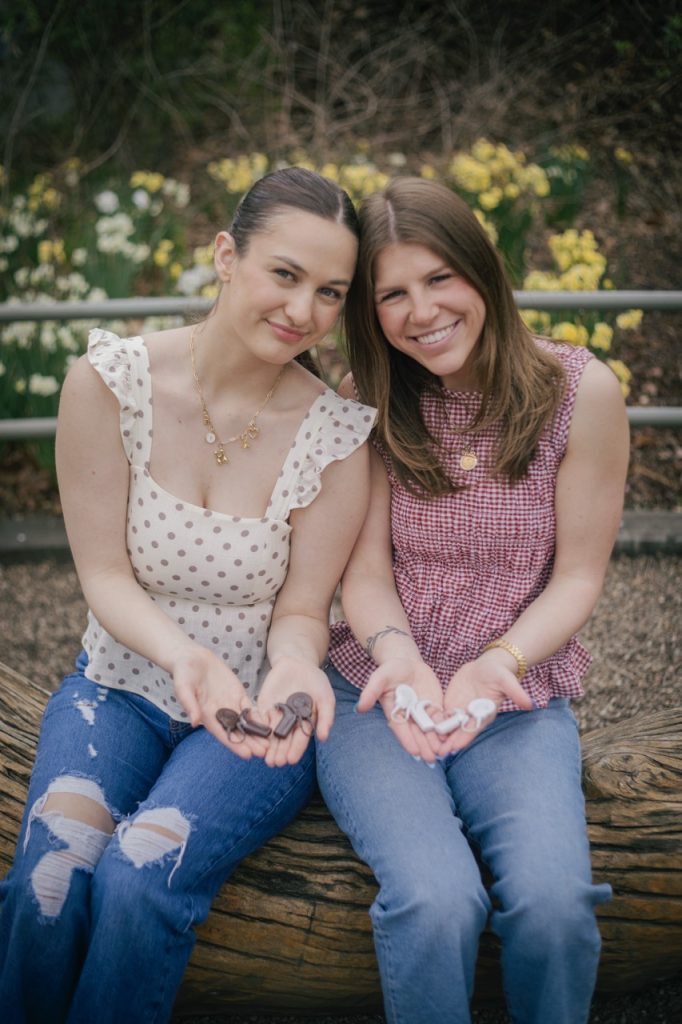 two girls sit smiling, holding cochlear implants in their hands in the shape of a heart.
