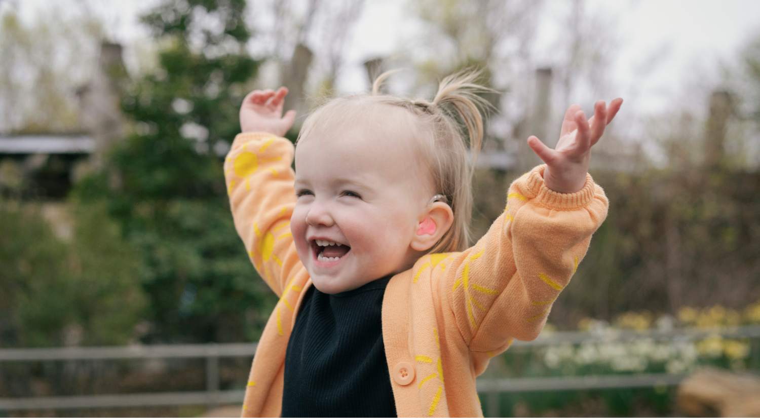 young girl with blonde hair and pink hearing aids is playing outside. She throws her hands in the air while laughing.