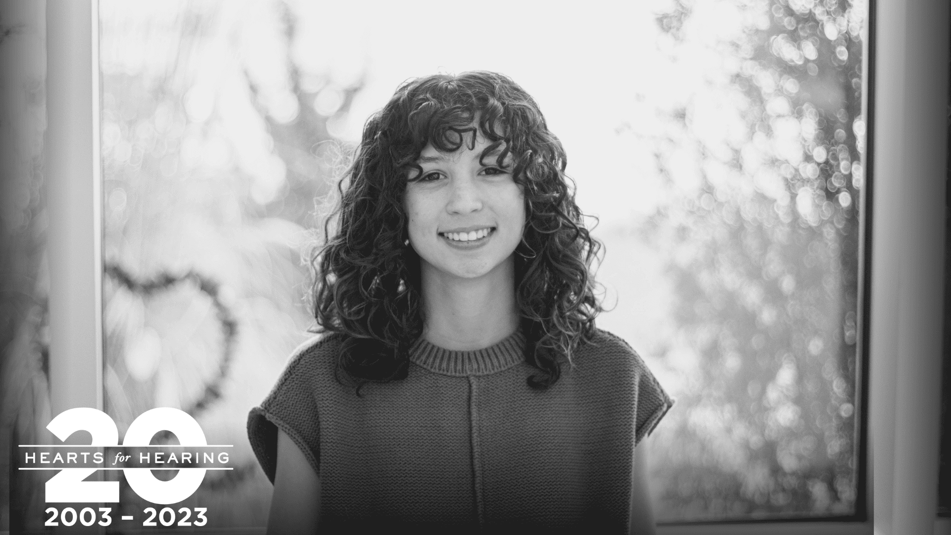a black and white photo of a girl with dark curly hair infront of a window
