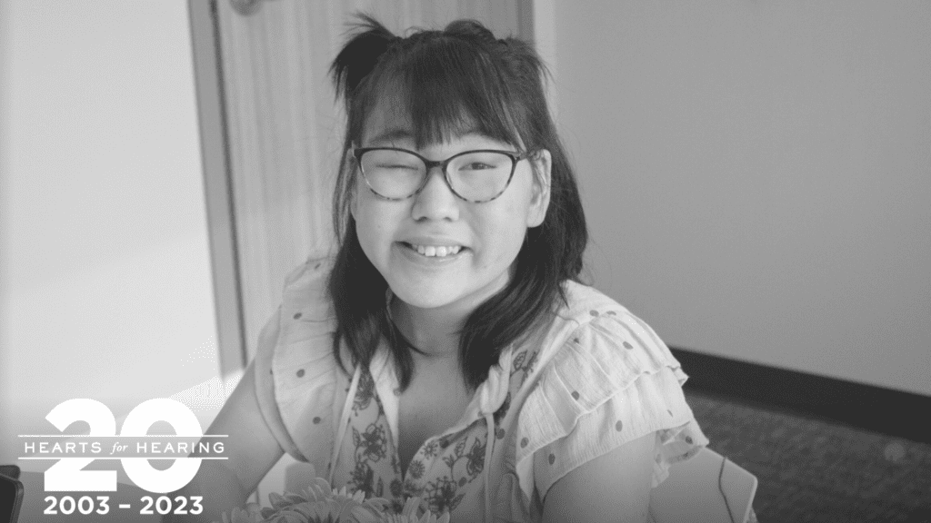 Photo of a girl with glasses smiling at the camera. She has dark hair and a light colored top.