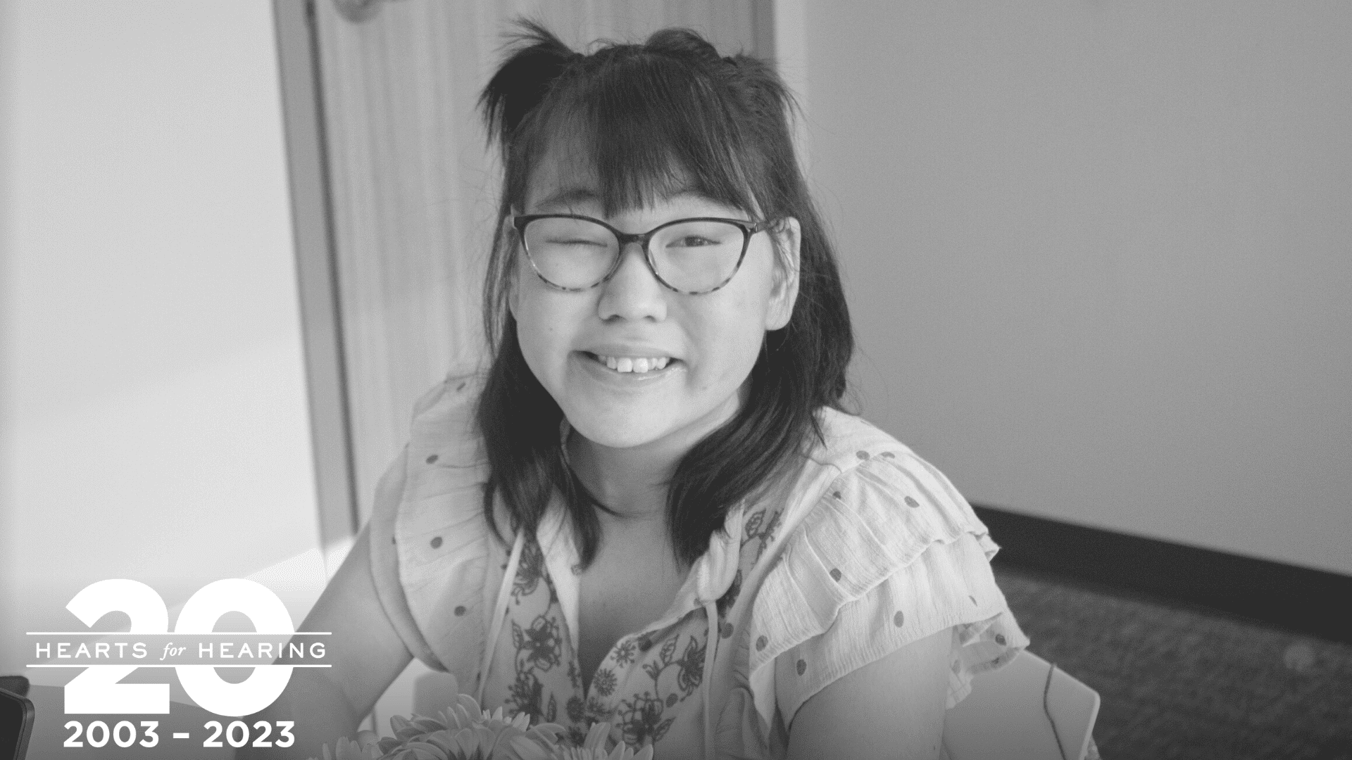 Photo of a girl with glasses smiling at the camera. She has dark hair and a light colored top.