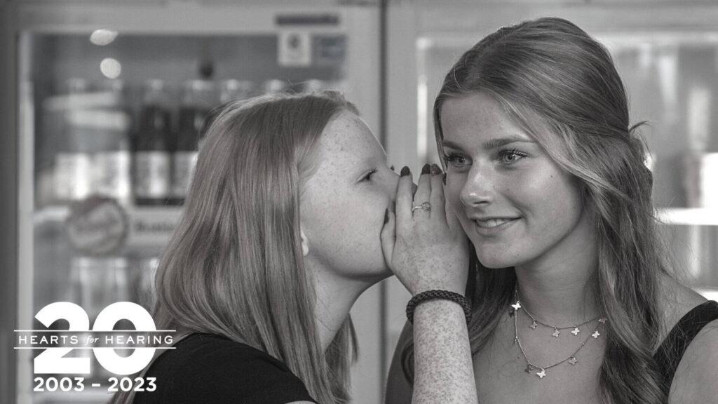 two girls stand in front of a fridge filled with soda. The girl on the left is leaning in to whisper a secret to the girl on the right.