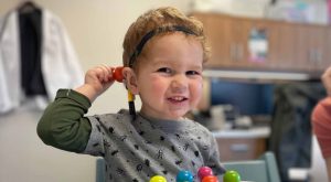 Boy grins as he hold a hold a small toy up to his cochlear implant