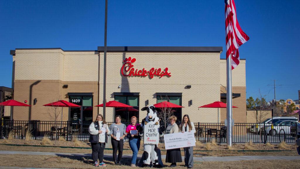 Five women stand around a mascot cow holding signs and a giant check infront of a tan brick building