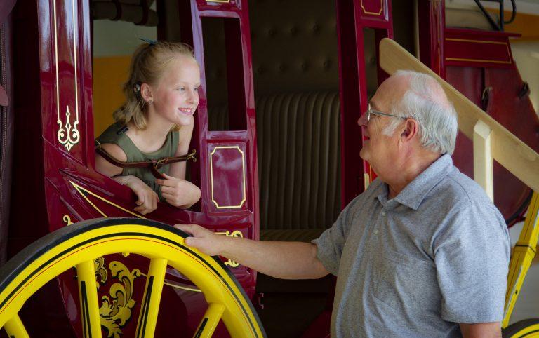 young girl sitting inside historic horse coach, grandfather smiling closely by