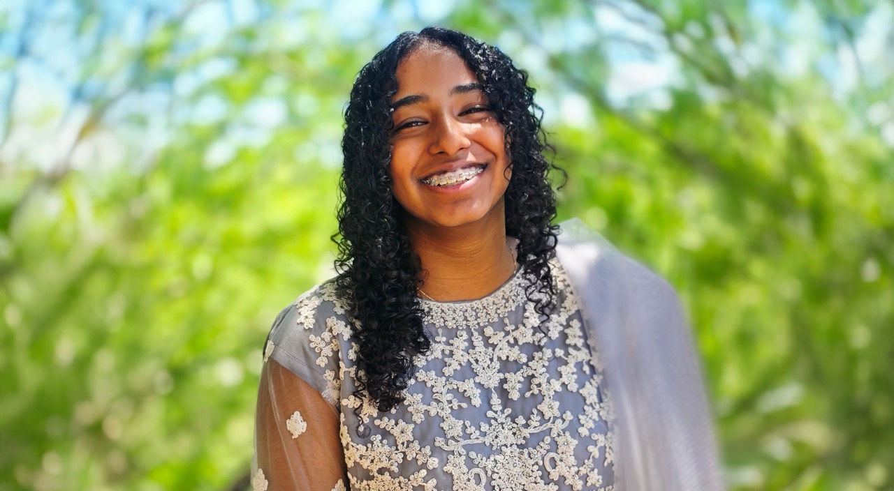 A girl with curly hairy and braces smiles at the camera, she is wearing a sari that is light purple with cream details