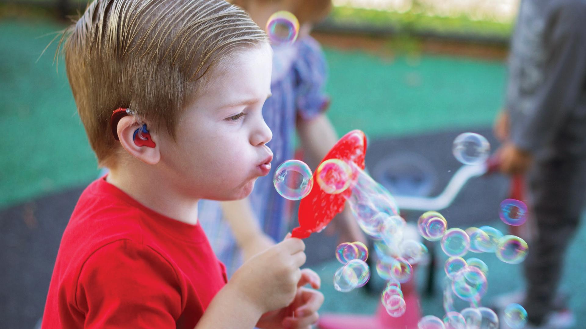 young boy in red shirt happily blowing bubbles and wearing matching red hearing aid devices