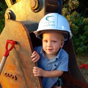 young smiling boy wearing hearts for hearing branded hard hat while sitting inside bulldozer bucket